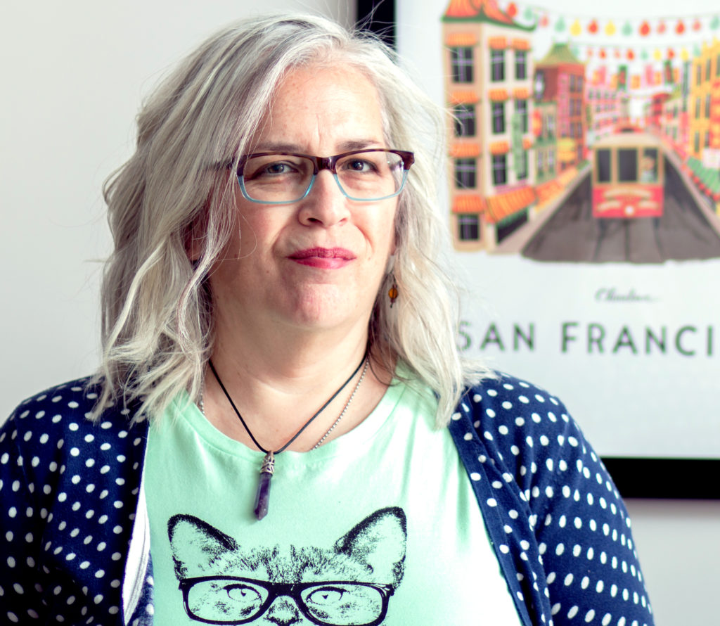 a headshot of Victoria Klum, a woman with white hair wearing glasses, a green t-shirt with a glasses-wearing tabby cat, and a blue sweater with white polka dots